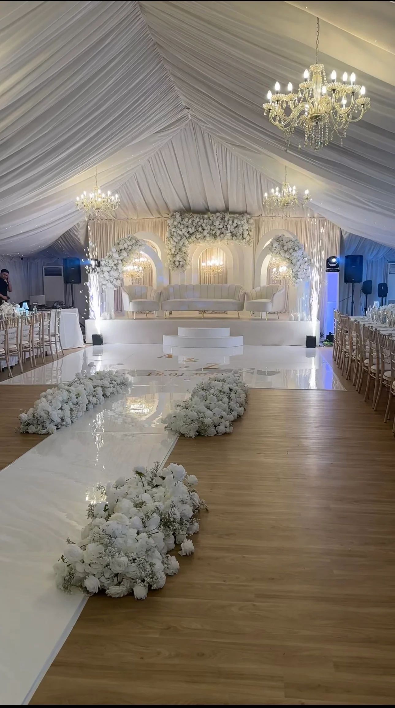 A seamless white dancefloor and aisle lined with white flowers in a draped marquee